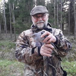 Pruunilaigulises riietuses, halli soni, habeme ja prillidega mees seisab metsa servas ja hoiab kahe käega halli värvi kägu.