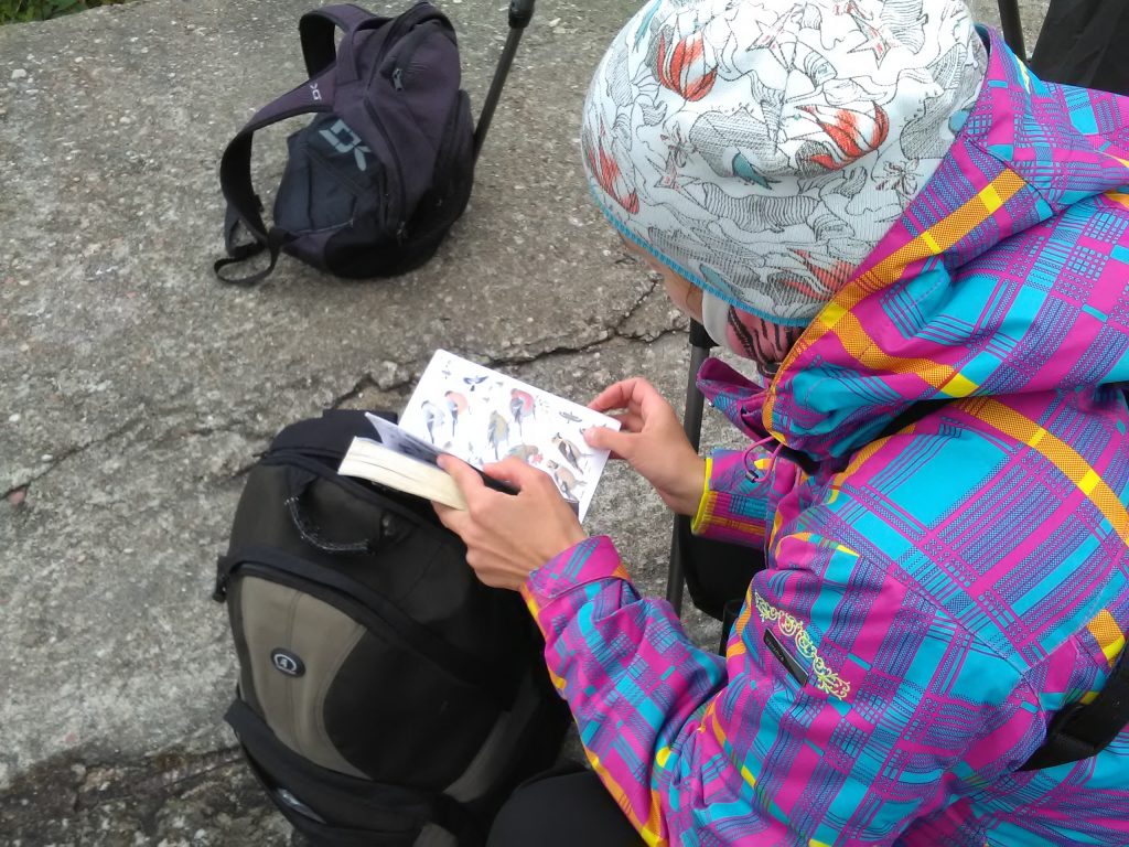 Kirjus jopes naisterahvas kükitab, tema ees maas on seljakott. Naine hoiab käes avatud linnumäärajat.